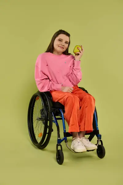 A young woman in a wheelchair sits in a studio setting, holding an apple and looking directly at the camera. She is wearing casual clothing and a smile. — Stock Photo