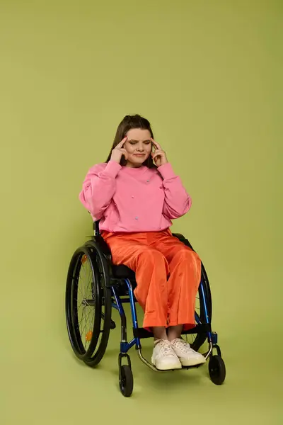 A young brunette woman in a wheelchair sits in a studio setting with a green background. She wears a pink sweater and orange pants. — Stock Photo