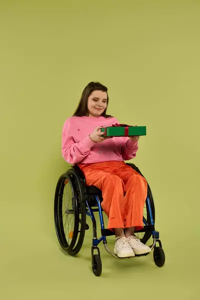 A brunette woman sits in a wheelchair against a green backdrop, smiling and looking at a wrapped gift shes holding. — Stock Photo