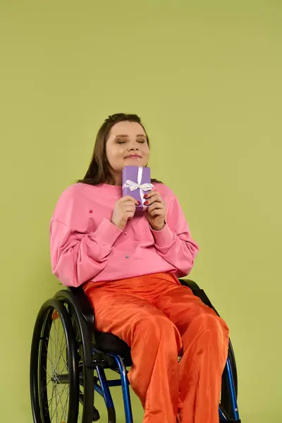 A young woman with dark hair sits in a wheelchair and smiles while holding a small gift box in her hands. — Stock Photo