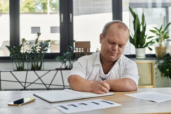 Ein inklusiver Mann sitzt an einem Schreibtisch in einem Büro und schreibt konzentriert in ein Notizbuch. — Stockfoto
