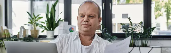 Человек с инклюзивностью работает над ноутбуком в современной офисной обстановке. Он окружен растениями и естественным светом. — стоковое фото