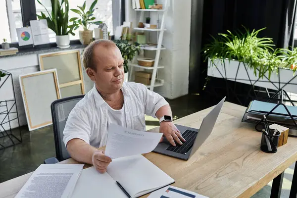 Um homem com inclusão senta-se em uma mesa em um escritório, olhando para um documento enquanto usa um laptop. — Fotografia de Stock