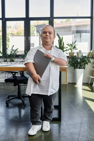 A man with inclusivity stands confidently in a modern office, holding a laptop, showcasing diversity and inclusion in the workplace. — Stock Photo