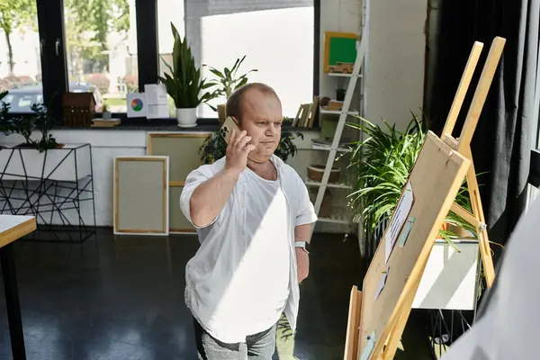 Un hombre con inclusividad habla por teléfono mientras está de pie en una oficina con una gran ventana y un caballete. - foto de stock