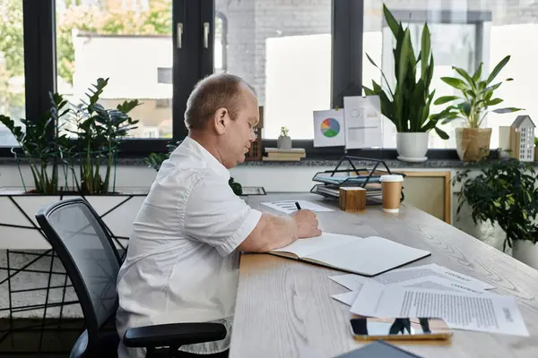 A man with inclusivity sits at a desk in a bright office, concentrating on writing in a notebook. — Stock Photo