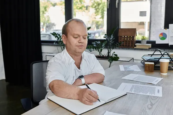 A man with inclusivity sits at a desk in an office, focused on writing in a notebook. — Stock Photo