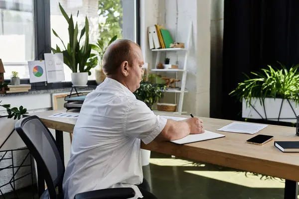 A man with inclusivity in a white shirt is seated at a desk in an office, intently focused on writing in a notebook. — Stock Photo
