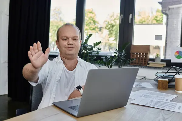 Un uomo con inclusività si siede a una scrivania in un ufficio, salutando la telecamera durante una videochiamata. — Foto stock