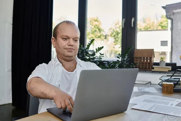 Un hombre con inclusividad se sienta en un escritorio en su oficina, trabajando intensamente en su computadora portátil. - foto de stock