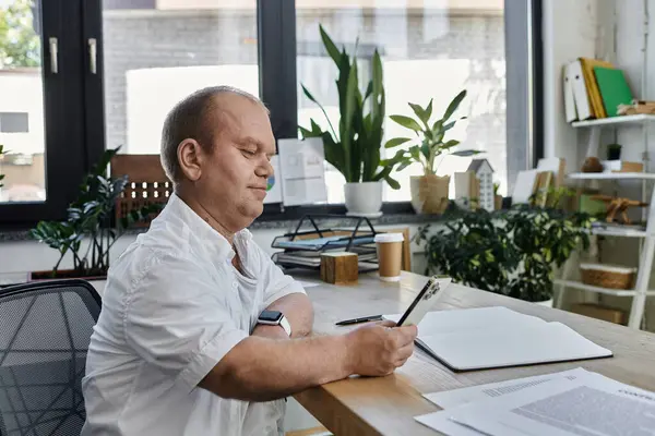 Um homem com inclusão senta-se em uma mesa em um ambiente de escritório, trabalhando em papelada com telefone. — Fotografia de Stock