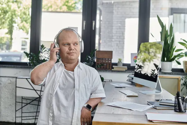 Un hombre con inclusividad usando auriculares se sienta en un escritorio en una oficina, escuchando música. - foto de stock
