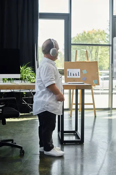 A man with inclusivity in a white shirt stands in an office, looking at a board, with headphones on. — Stock Photo