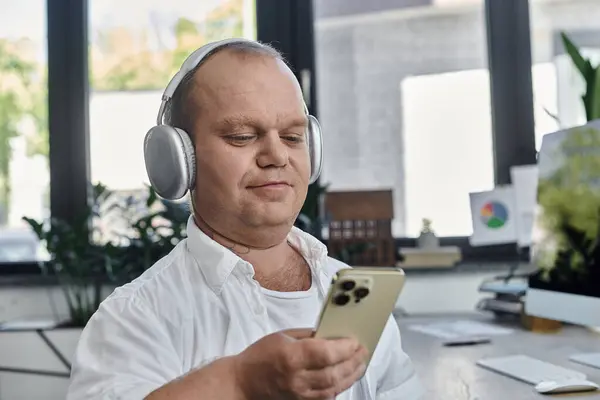 A man with inclusivity wearing headphones and looking at his phone, taking a moment to relax in a busy office. — Stock Photo