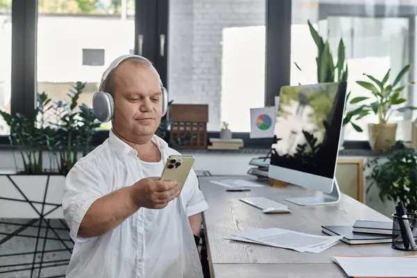 Человек с инклюзивностью в наушниках сидит за своим столом в офисе, используя свой смартфон. — стоковое фото