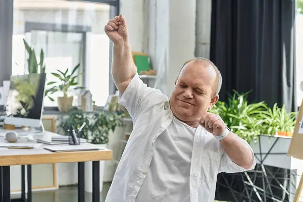 A man with inclusivity in a white shirt stretches his arms overhead while standing in an office setting. — Stock Photo