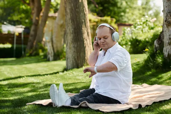 A man with inclusivity sits on a blanket in a park, wearing headphones and a smartwatch. — Stock Photo