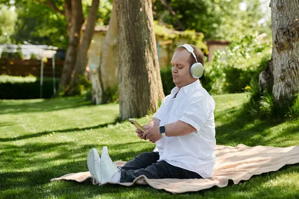 A man with inclusivity sits on a blanket in a grassy park, wearing headphones and looking at his phone. — Stock Photo