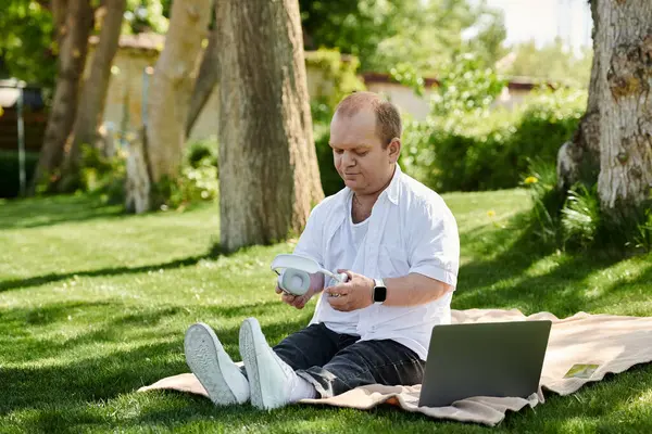 A man with inclusivity sits on a blanket in a park, surrounded by green grass, and puts on his headphones. — Stock Photo
