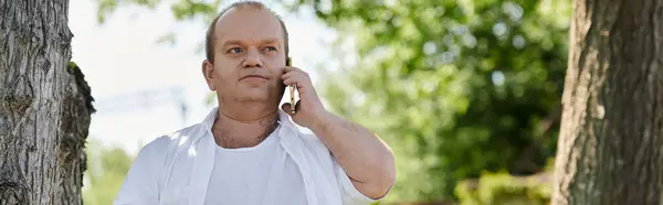 A man with inclusivity in a white shirt stands near a tree, looking contemplative as he speaks on his phone. — Stock Photo