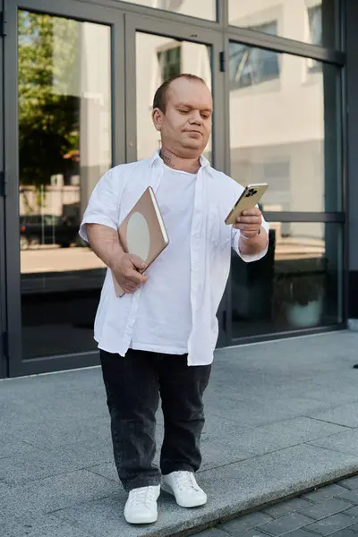 A man with inclusivity in a white shirt stands outside a building, holding a notebook and a smartphone. — Stock Photo