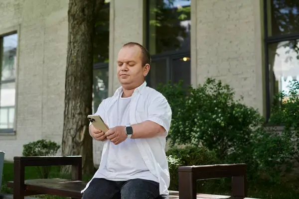 A man with inclusivity sits on a bench, engrossed in his phone, amidst the greenery of a city park. — Stock Photo