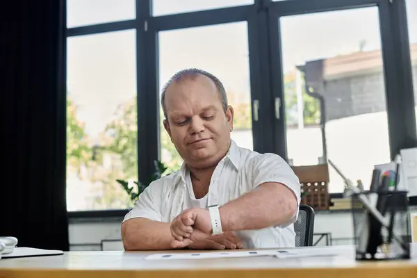 Человек с включённостью в белой рубашке проверяет время на наручных часах, сидя за столом в офисе. — стоковое фото