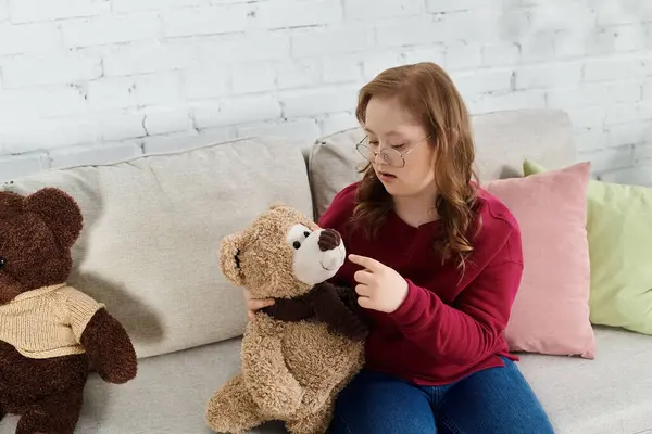 Girl with Down syndrome cuddles teddy bears on couch, cherishing childhood joys. — Stock Photo