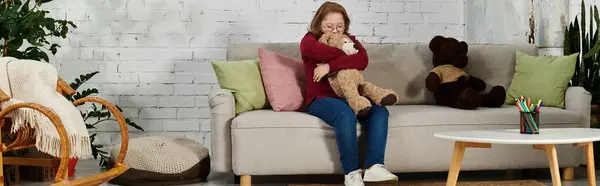 Uma menina com síndrome de Down senta-se em um sofá, segurando um urso de pelúcia perto. — Fotografia de Stock