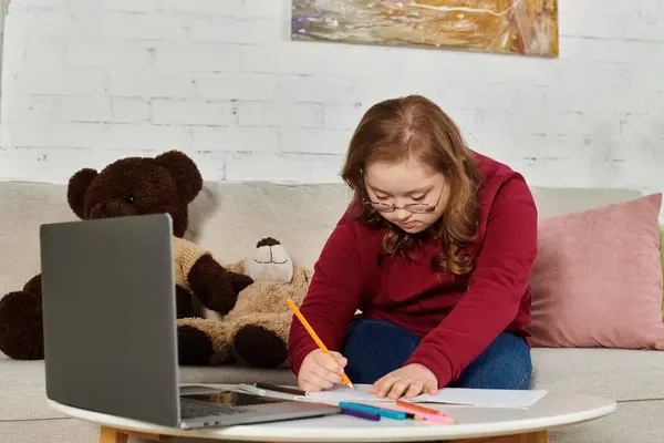 A little girl with Down syndrome sits on a couch, drawing at home. — Stock Photo