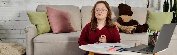 Ein kleines Mädchen mit Down-Syndrom sitzt an einem Tisch in ihrem Haus und lernt mit Notizbuch und Laptop. — Stockfoto