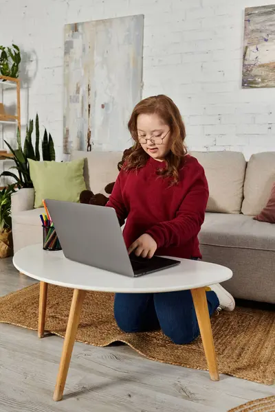 A little girl with Down syndrome sits on the floor, using a laptop at a coffee table in her home. — Stock Photo