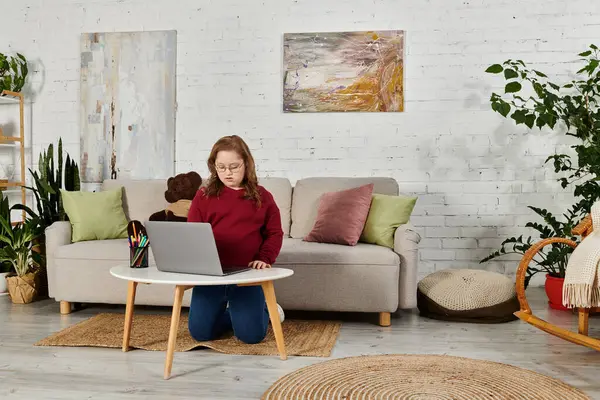 A little girl with Down syndrome uses a laptop while sitting on the floor in her living room. — Stock Photo