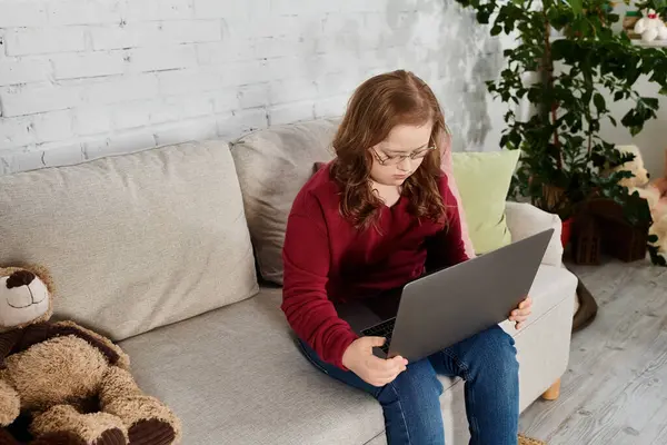A little girl with Down syndrome sits on a couch at home, using a laptop. — Stock Photo