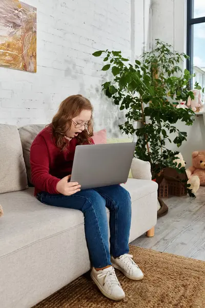 A little girl with Down syndrome sits on a couch at home, surprised by something on her laptop. — Stock Photo