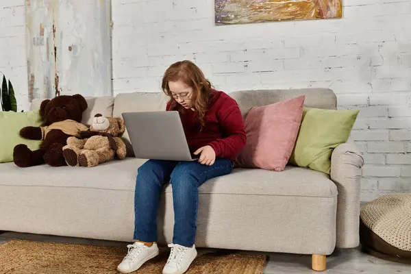 A little girl with Down syndrome sits on a couch at home, using a laptop. — Stock Photo