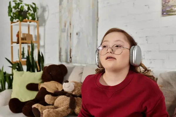 A little girl with Down syndrome wearing headphones, sitting on a couch in a home setting. — Stock Photo