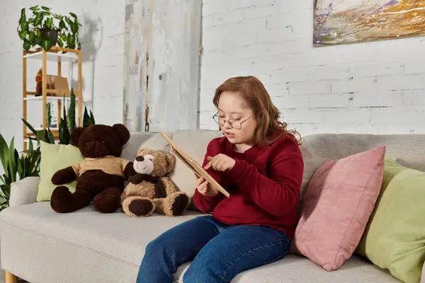 A little girl with Down syndrome sits on a couch, surrounded by plush toys, reading a book. — Stock Photo