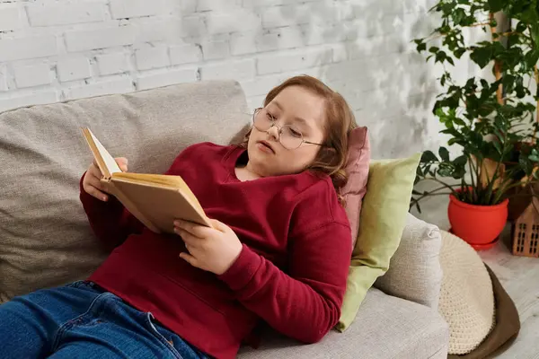 A little girl with Down syndrome relaxes on a couch at home, engrossed in a book. — Stock Photo