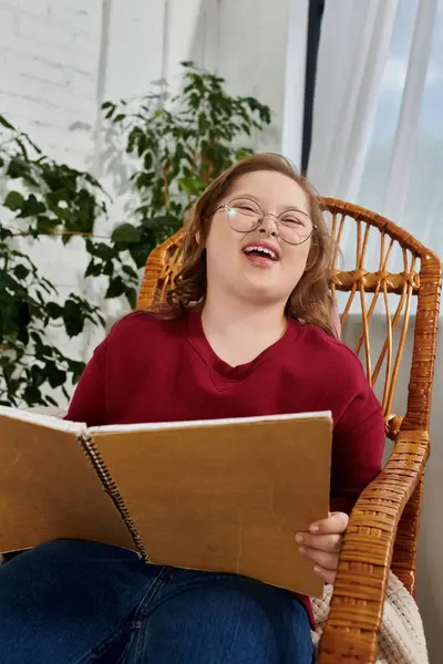 A little girl with Down syndrome laughs while reading a book in a wicker chair. — Stock Photo