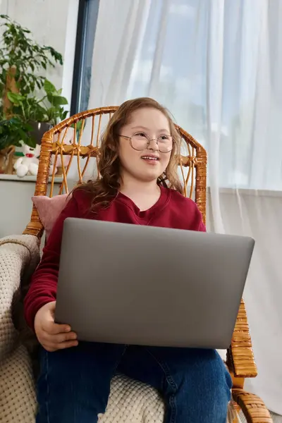 A little girl with Down syndrome uses a laptop while sitting in a wicker chair. — Stock Photo