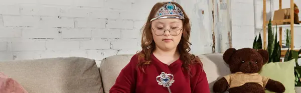 A little girl with Down syndrome sits on a couch, wearing a crown and holding a wand. — Stock Photo
