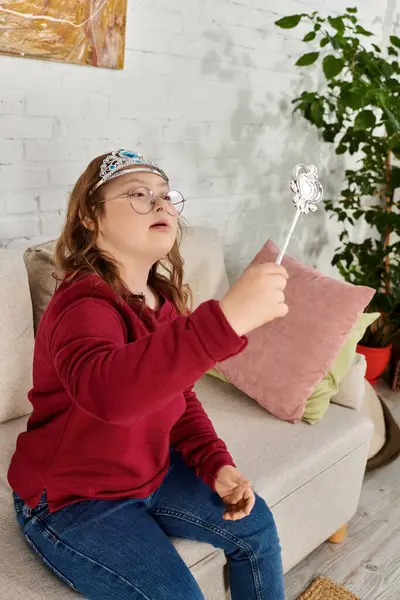 Girl with Down syndrome sits on couch, wearing crown and holding wand, lost in imagination. — Stock Photo