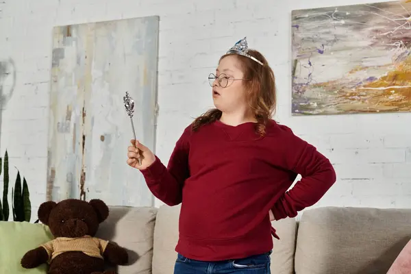 A little girl with Down syndrome plays with a toy wand while wearing a crown, at home. — Stock Photo