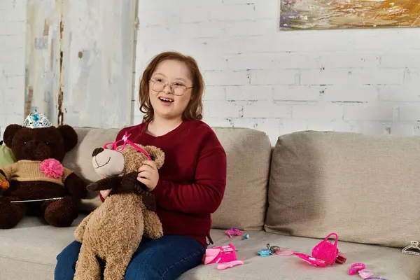 A little girl with Down syndrome sits on a couch, holding a teddy bear, and smiling brightly. — Stock Photo
