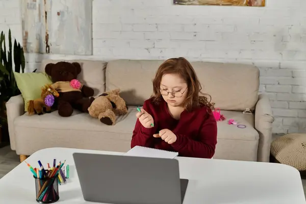Little girl with Down syndrome at desk, writing in notebook, with open laptop. — Stock Photo