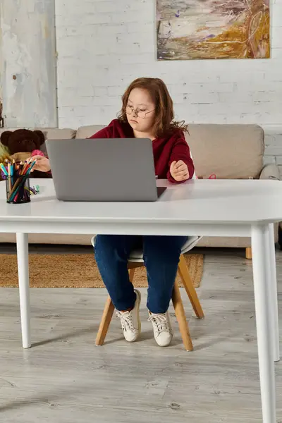 A little girl with Down syndrome sits at a table, focused on her laptop in a home setting. — Stock Photo