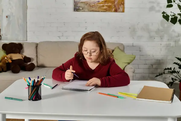 Una bambina con la sindrome di Down siede a un tavolo disegnando in un quaderno. — Foto stock