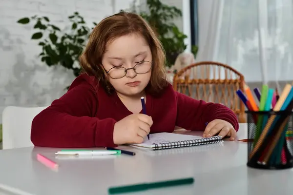 Una bambina con la sindrome di Down siede a un tavolo, disegnando intensamente nel suo taccuino.. — Foto stock