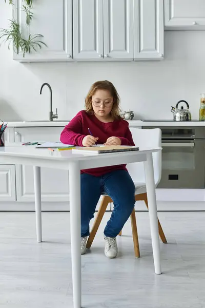 Ein kleines Mädchen mit Down-Syndrom sitzt am Küchentisch und zeichnet oder schreibt fleißig. — Stockfoto